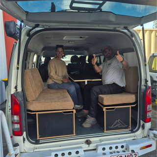 Two people sitting inside a van with converted seats and a table, giving thumbs up.